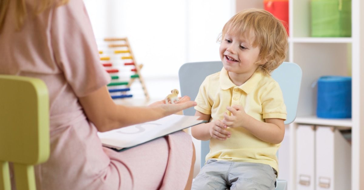 A therapist offers a small toy to a smiling young boy during a seated session in a bright, colorful playroom.