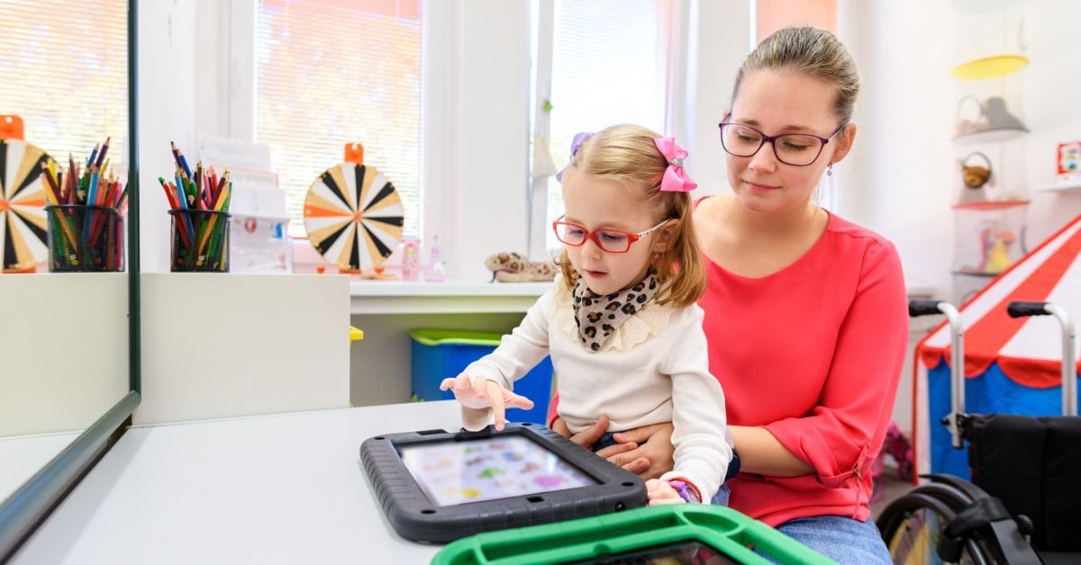 A young girl taps a tablet while a woman supports her in a bright therapy room filled with colorful 