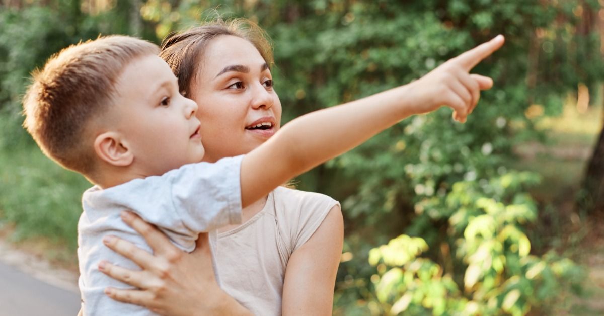 A young boy points into the distance while a woman holds him outdoors on a leafy path in the green woods.