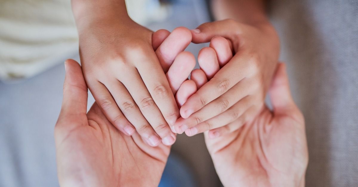 A child places both hands over an adult’s open hands in a close-up gesture of support and connection.
