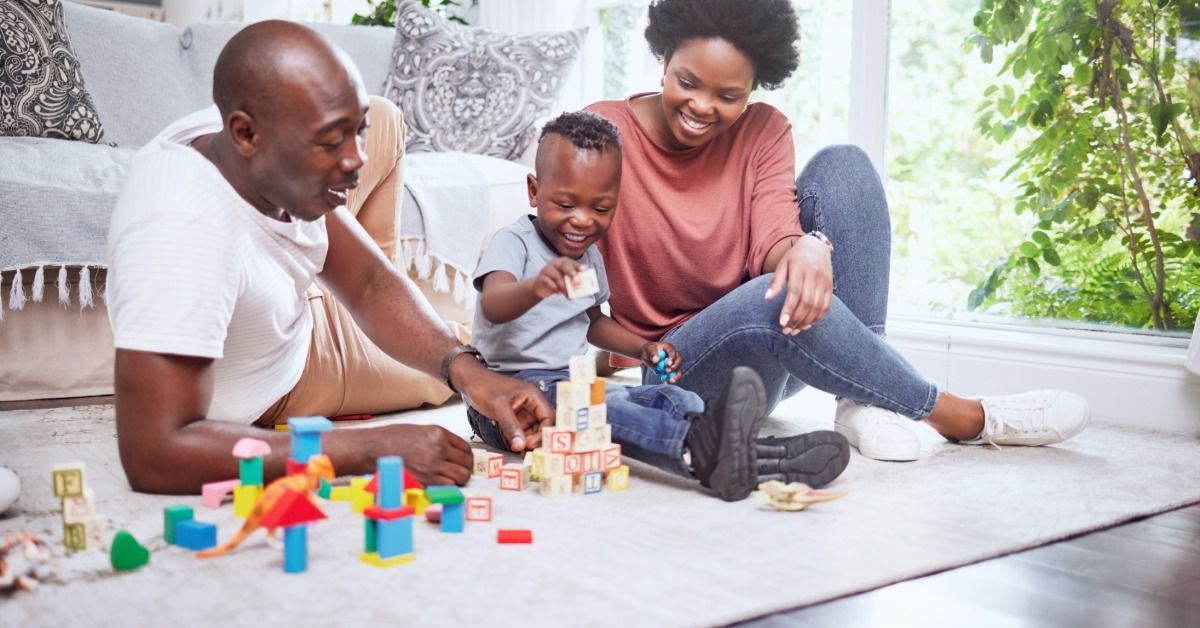A young boy stacks colorful blocks on the living room floor while his parents sit beside him.