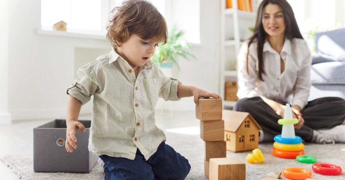 A young boy stacks multiple wooden blocks on a carpet while his mother watches him in the background.