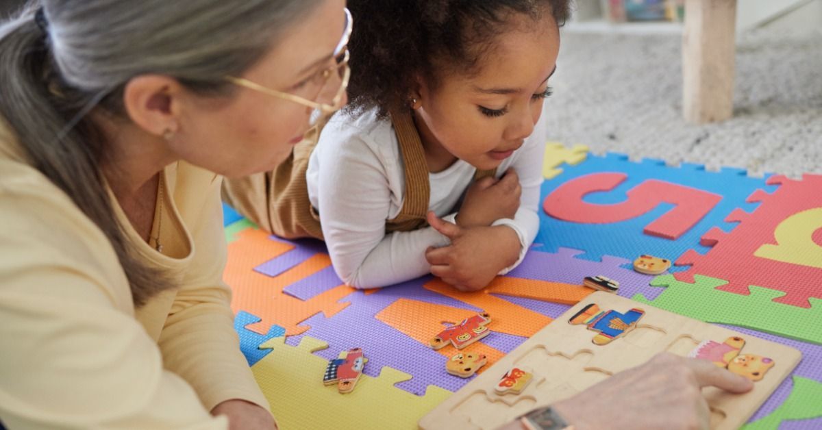 A young girl works on a wooden puzzle with a woman beside her as colorful foam letters cover the play mat.