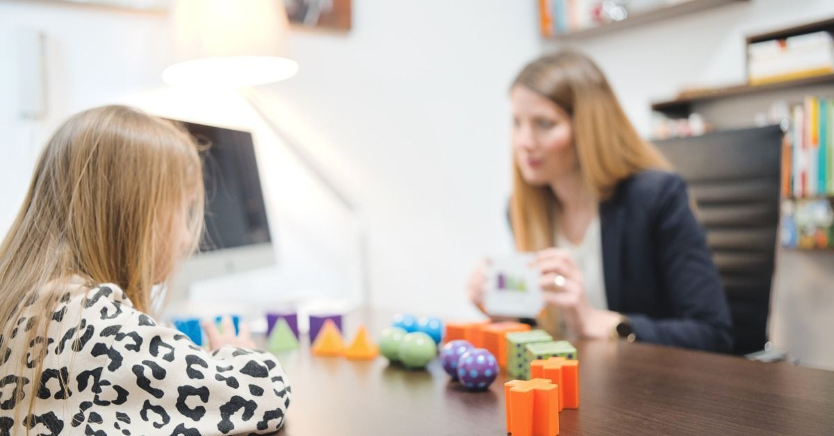 A therapist shows a card to a young girl seated across a table with colorful assessment toys spread out on it.