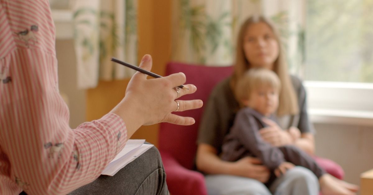 A professional gestures with a pencil while a mother holds her young child during an autism consultation.
