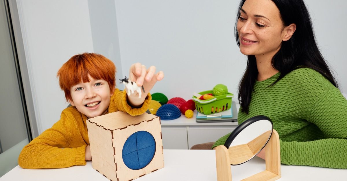A smiling boy holds a toy cow beside a woman as therapy toys and a tabletop mirror sit in front of them.