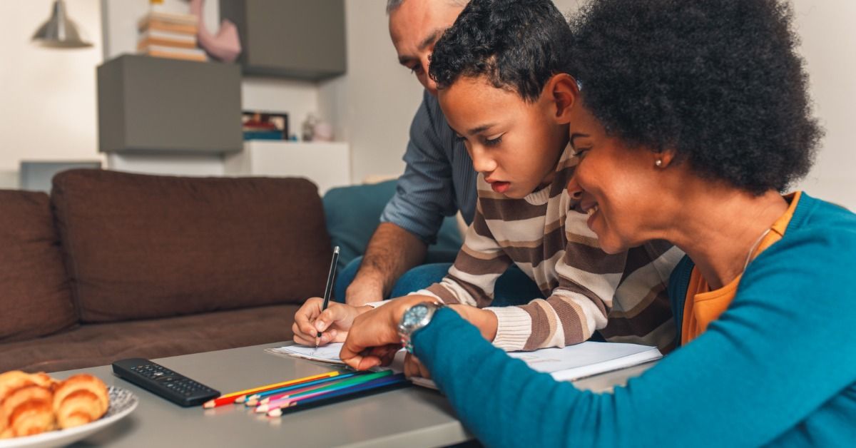 A mother and father help their child complete homework at a living room table.