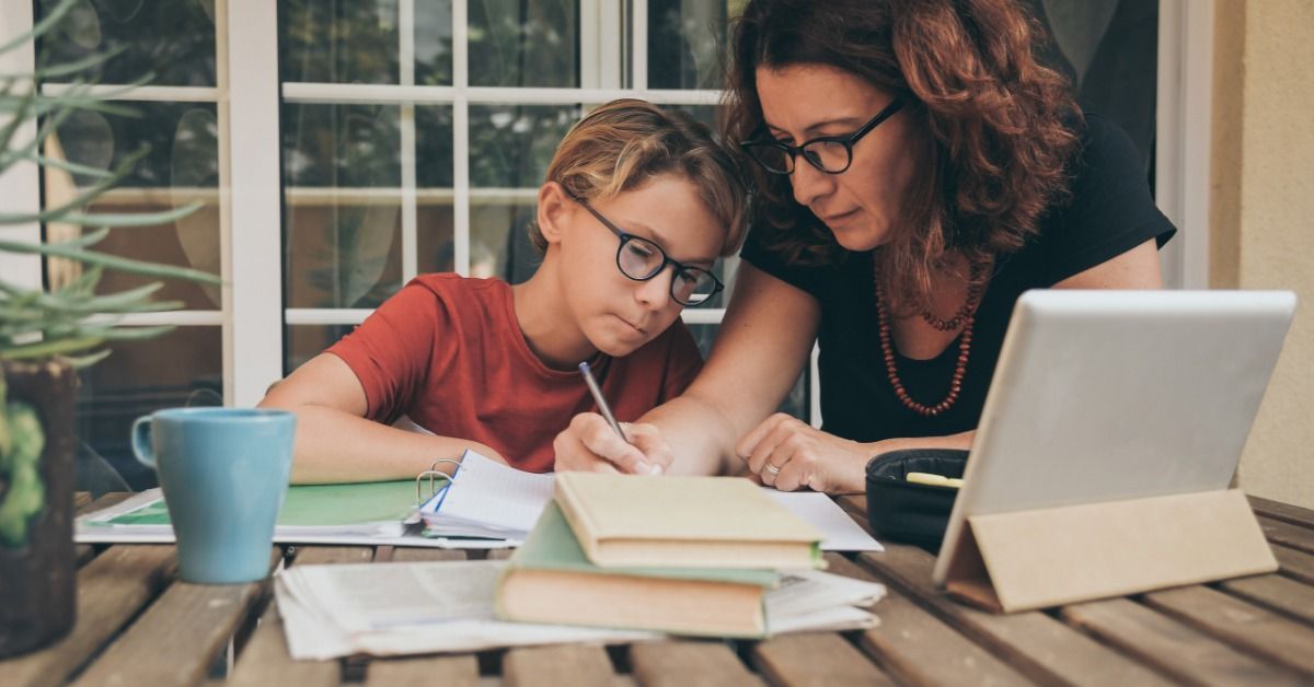 A mother helps her son complete homework at an outdoor table that has books, notebooks, and a tablet.