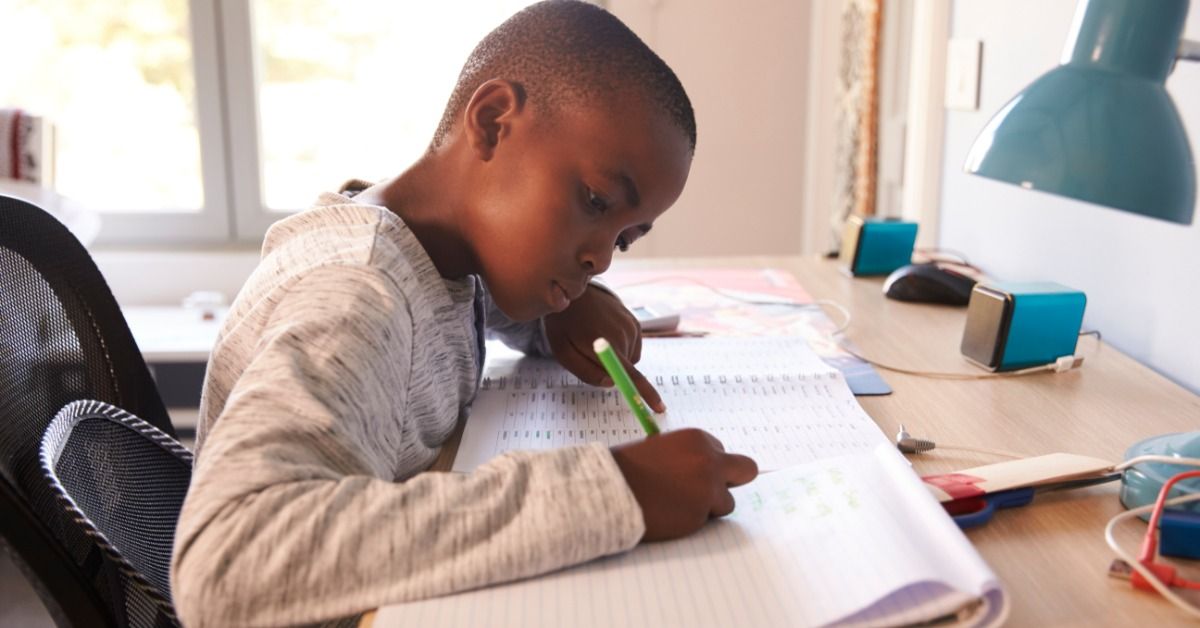 A young boy writes in a notebook at a bedroom desk while a lamp and school supplies sit nearby on the desk.