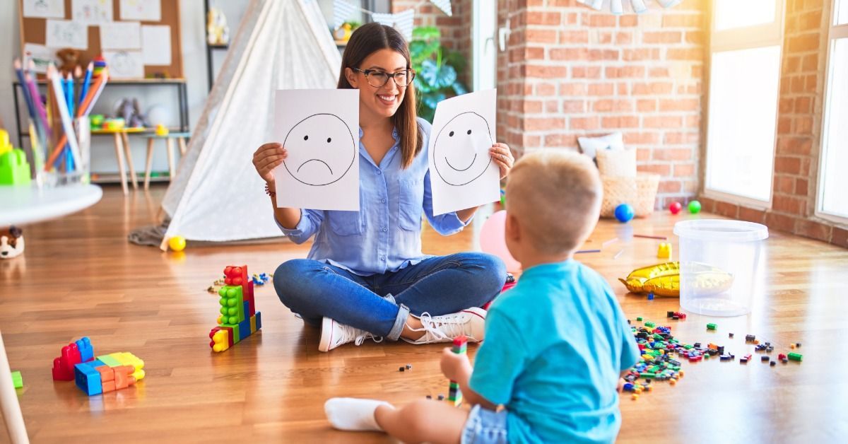 A therapist shows a young boy happy and sad face cards as they sit on a playroom floor.