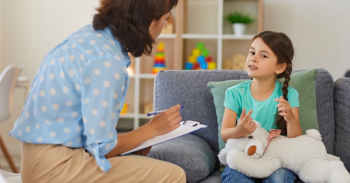 A young girl talks with a therapist while holding a stuffed bear on a couch in a bright, child-friendly therapy room.