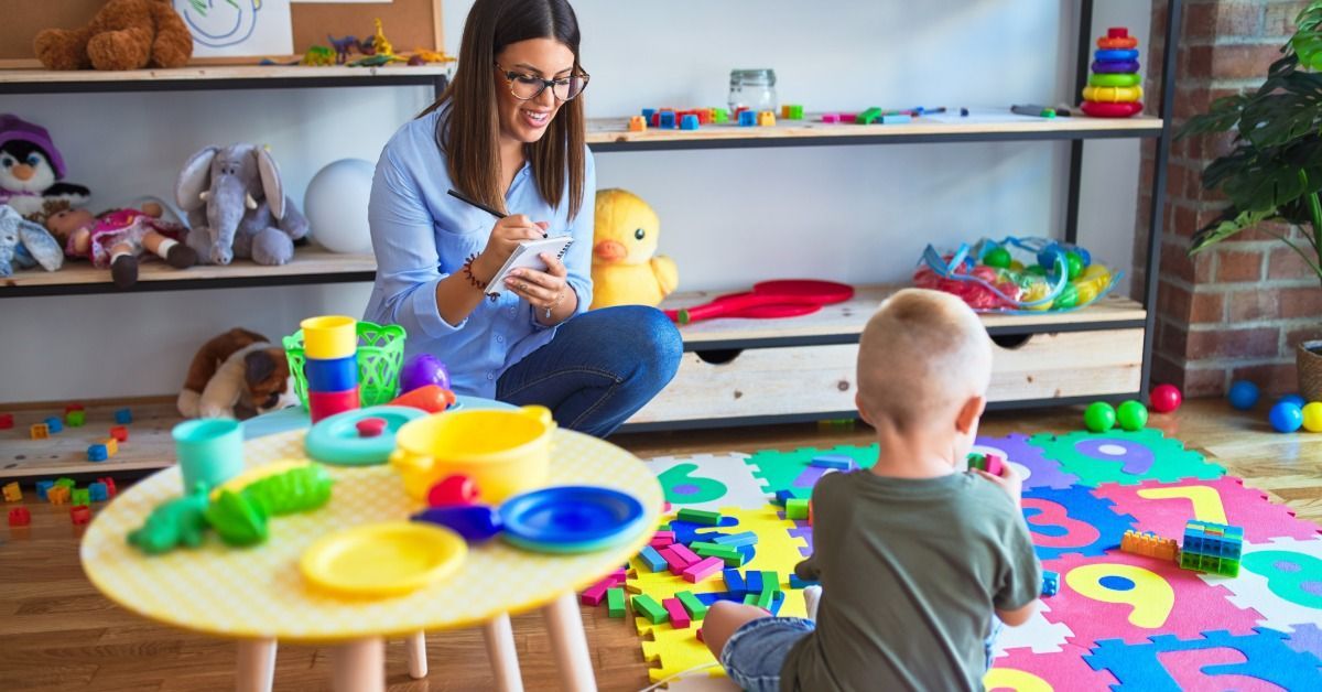 A therapist takes notes while a young boy plays with colorful blocks on a foam mat in a bright playroom.