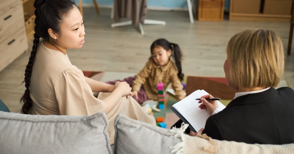 A mother talks with a therapist who takes notes while a young girl plays with colorful blocks.