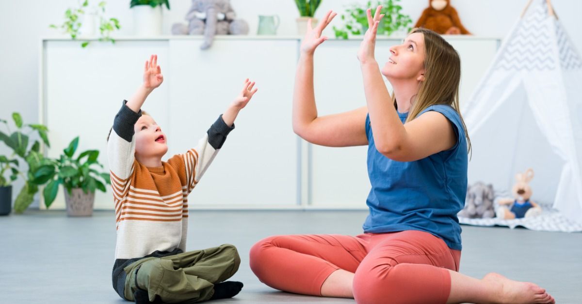 A young boy mirrors a therapist raising both hands as they sit together on the floor in a bright playroom.