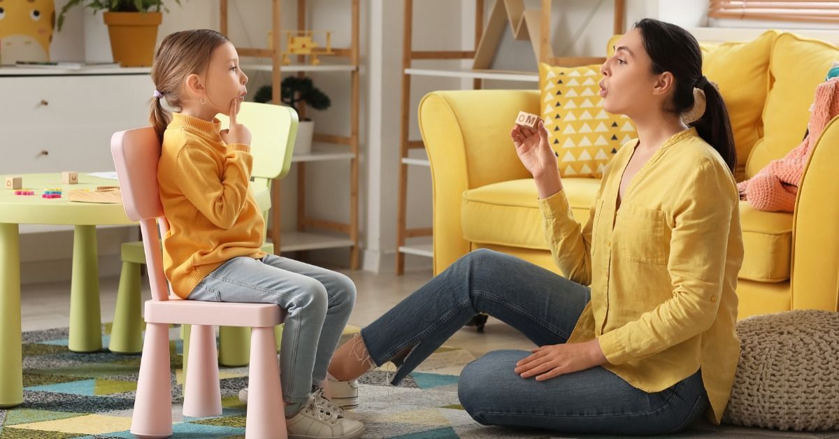 A speech therapist holds a letter block and models a sound as a young girl watches from a small chair.