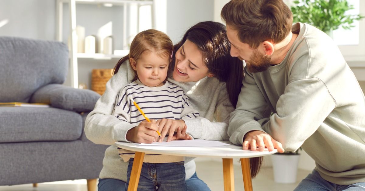 A mother guides a toddler's hand as the child draws at a small table while a father watches nearby.