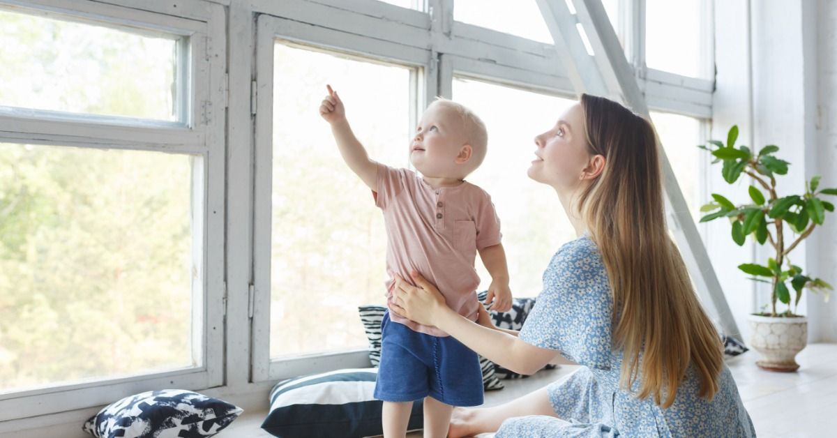 A mother helps her toddler stand beside a large window as the child points outside and they look out the window together.