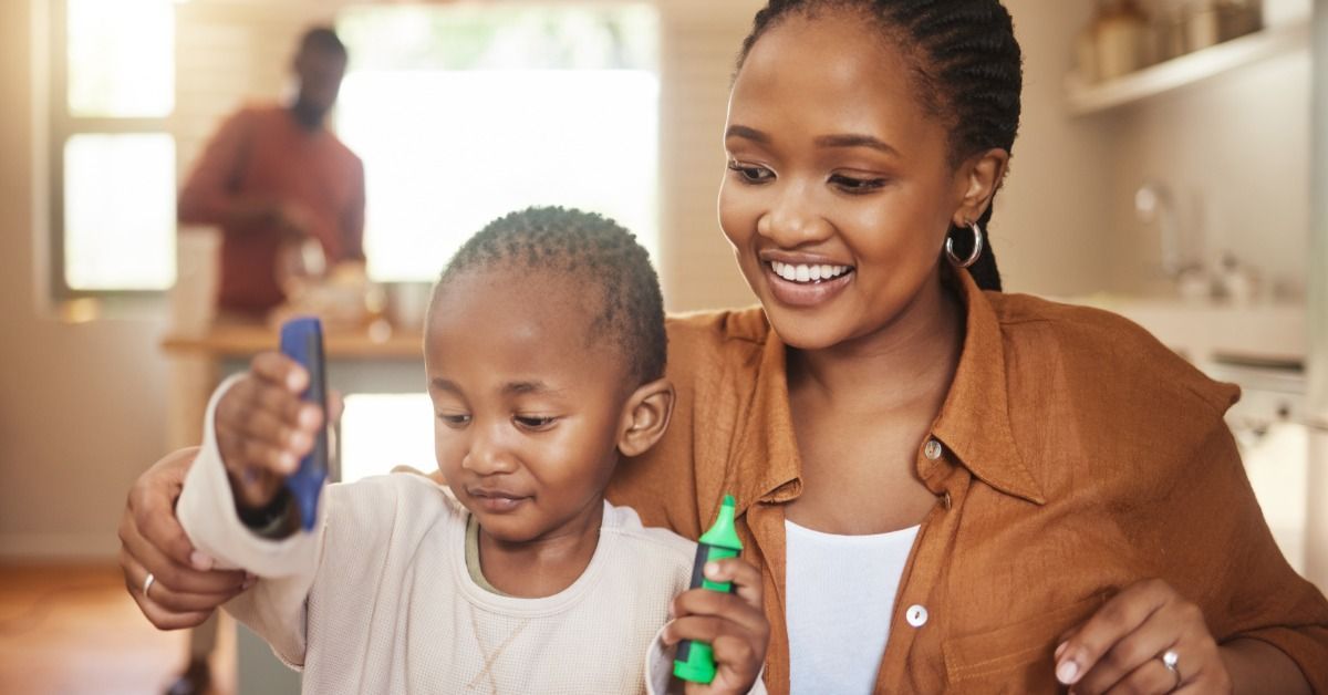 A mother smiles beside a young boy as he draws at a table with markers while a father stands in the background.