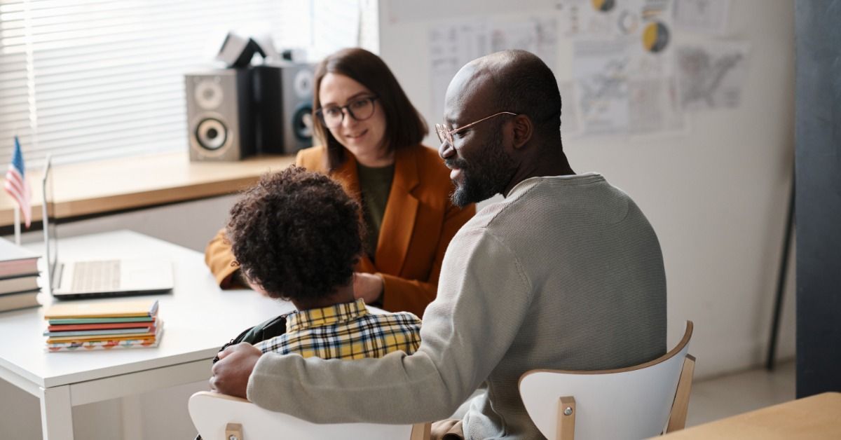 A father puts his arm around his son as they meet with a teacher at a desk.