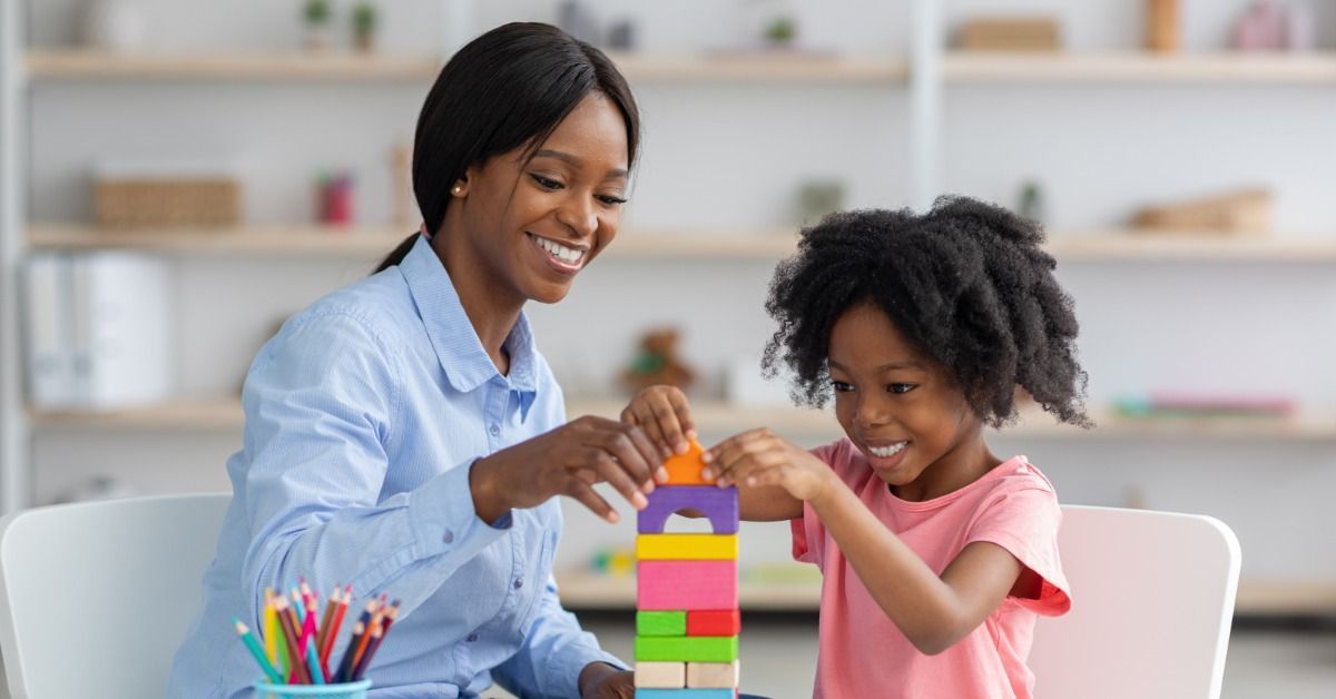 A woman and child stacking colorful blocks together at a table with pencils and papers in a bright room.