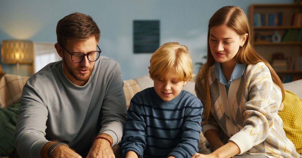 A man, woman, and child making crafts with paper, glue, and small objects at a table in a living room.