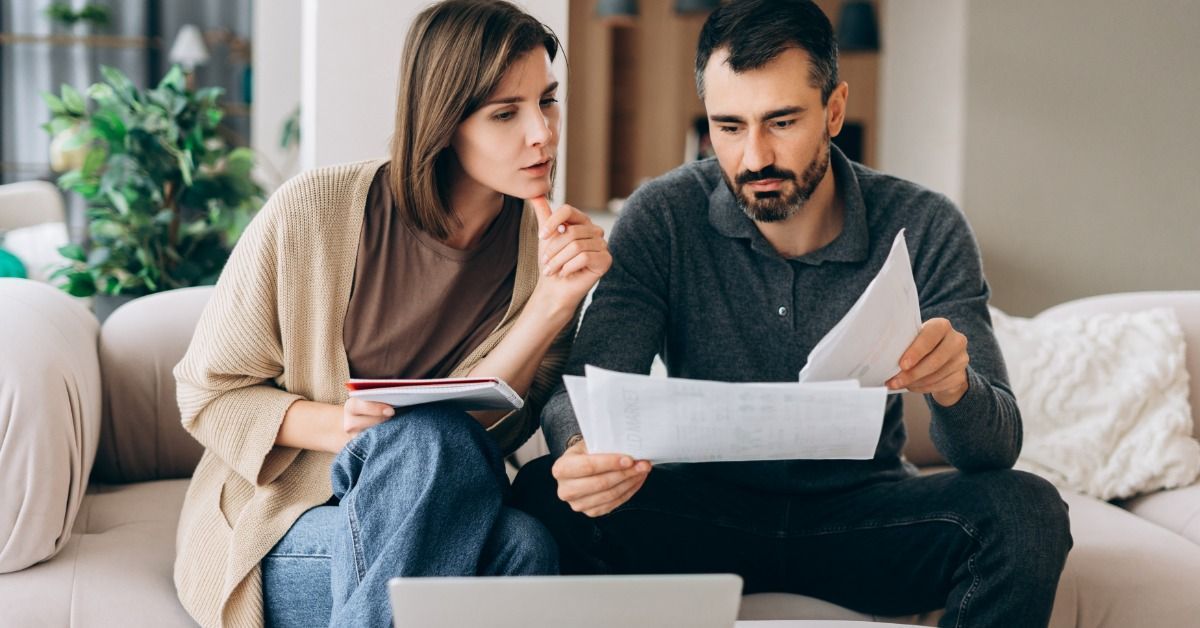 A woman and a man sitting on a sofa reviewing paperwork together, with a laptop on the table in fron
