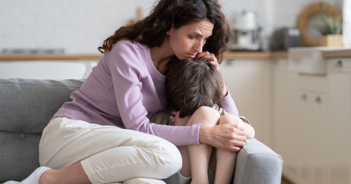 A woman in a purple sweater hugging a young boy curled up on a gray sofa, resting her hand on him.