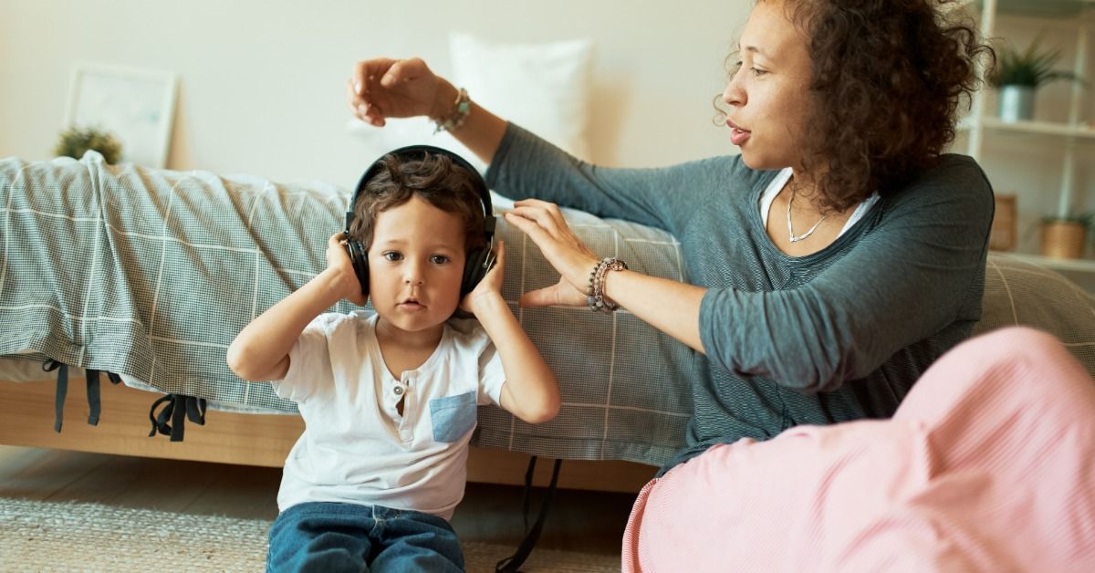 A young boy wearing black headphones and sitting on the floor while a woman beside him reaches toward his head.