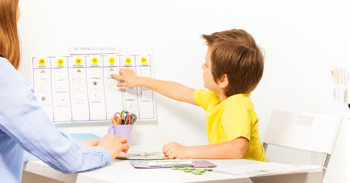 A boy in a yellow shirt pointing to a weekly calendar on the wall while sitting across a table from a woman.