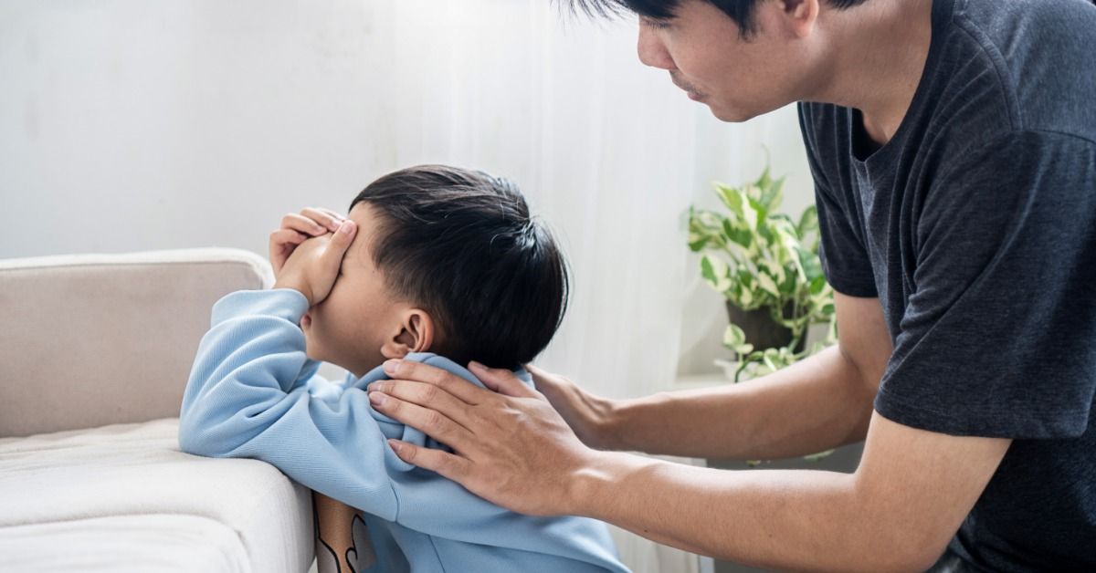 A young boy in a blue sweatshirt covering his face beside a couch while a man leans over to comfort.
