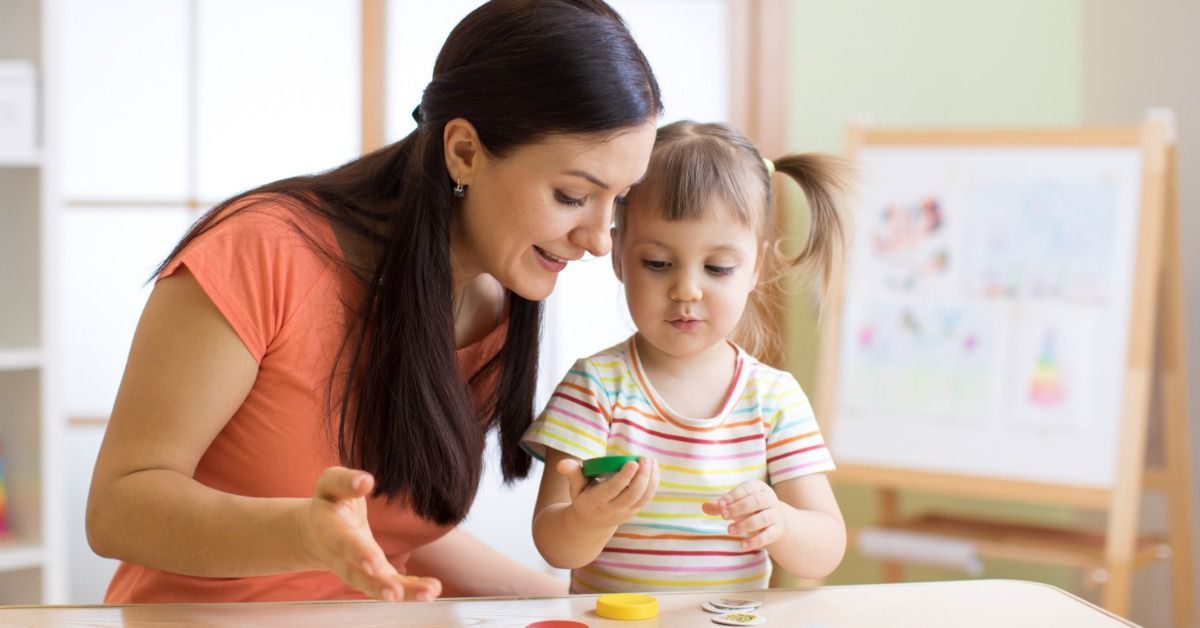 A woman in a coral shirt leaning toward a young girl holding a green game piece at a table covered with colorful pieces.
