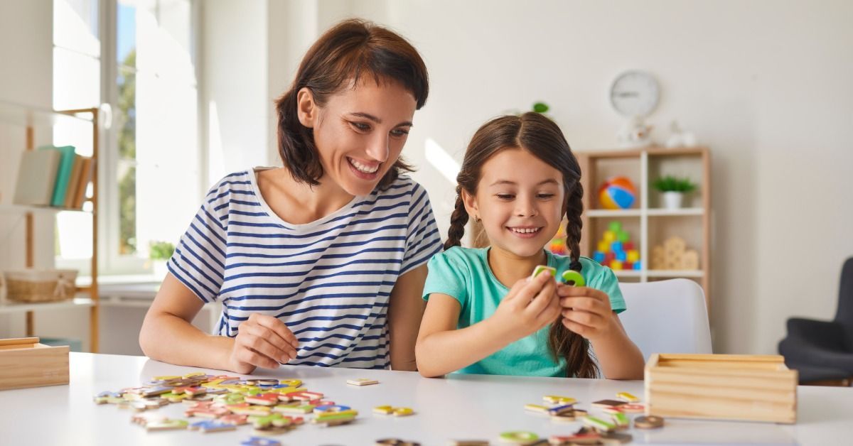 A smiling woman and young girl sitting together at a white table, handling colorful puzzle pieces spread across the surface.