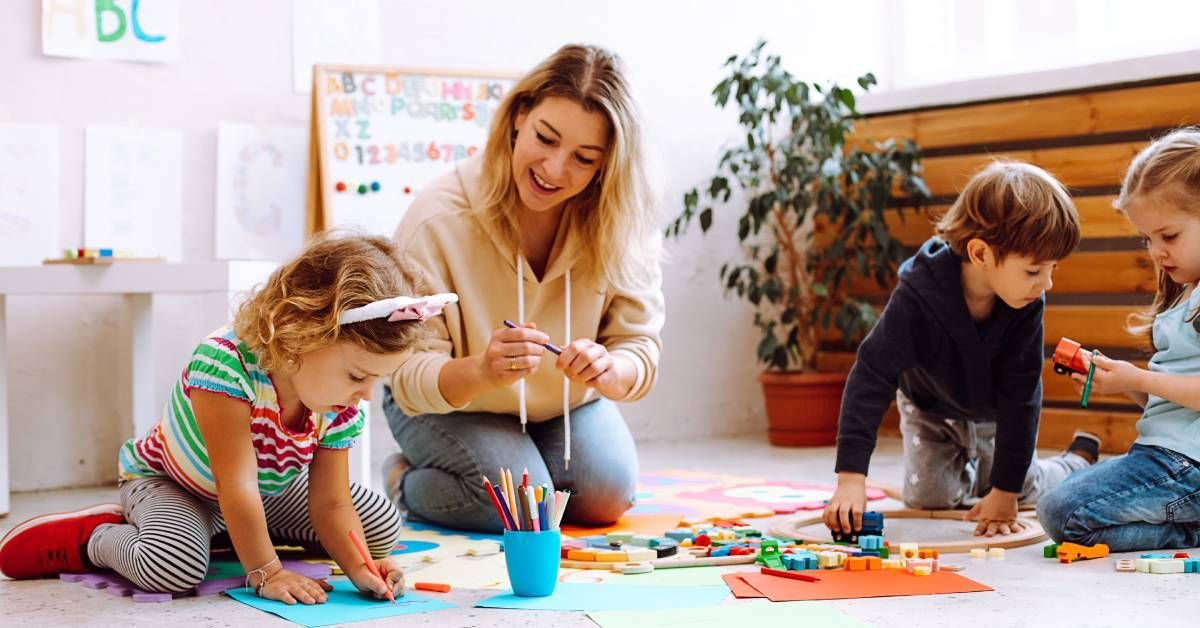 A young teacher smiling as she plays with a group of children who play on the floor with toys and arts and crafts.