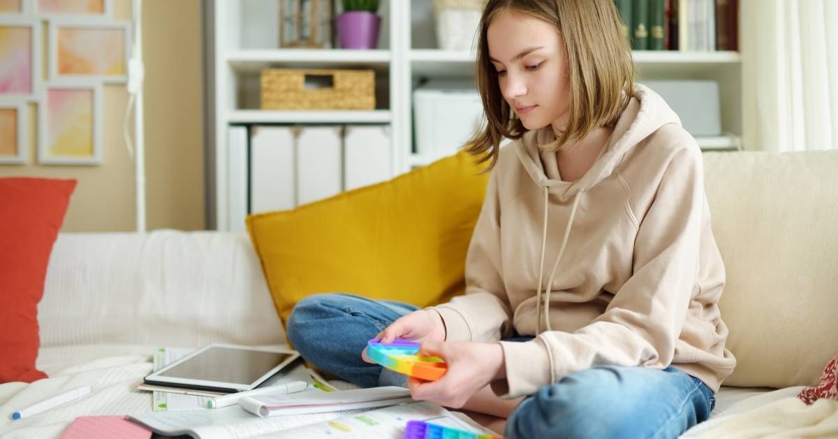 A teen girl is playing with a fidget sensory toy on the couch while doing homework.