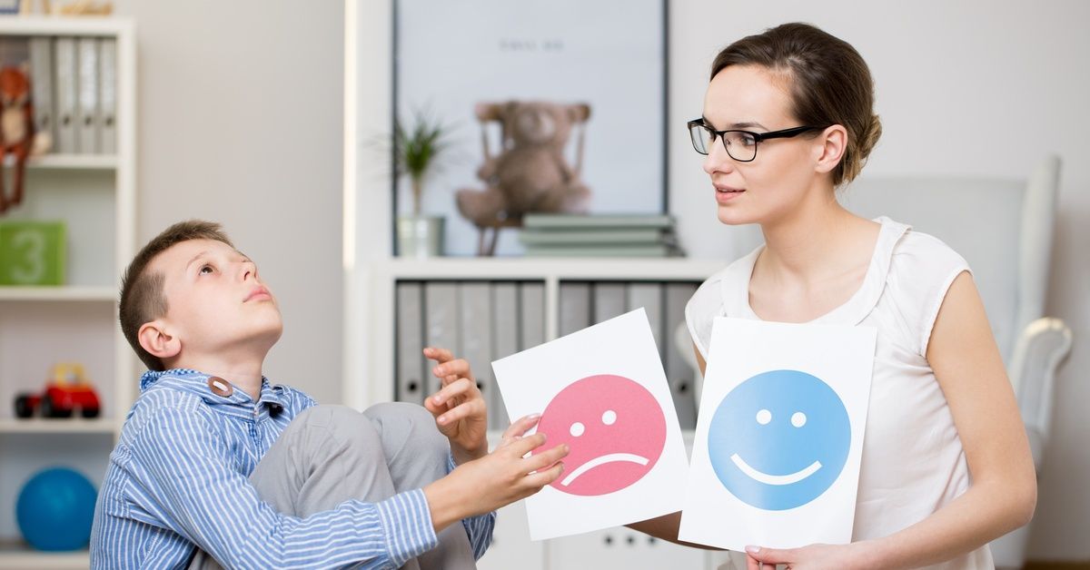 A little boy pointing to a sad face on a card while sitting near a woman who is holding up two cards.