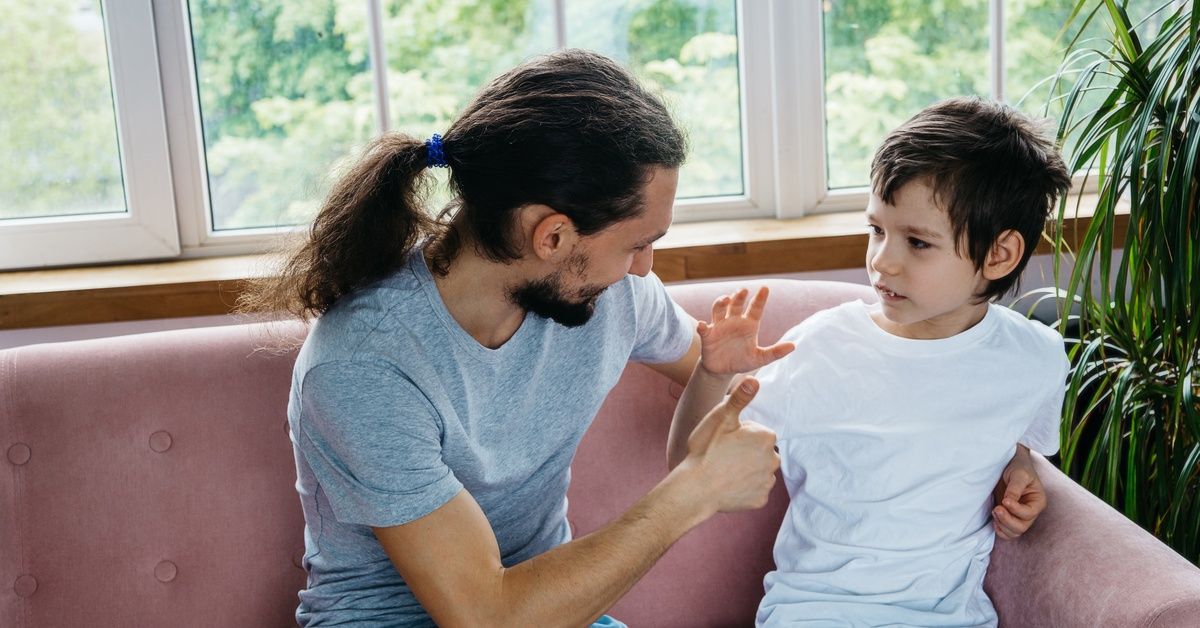 A man and a little boy sitting together on a couch as the man makes a thumbs-up sign toward the child.