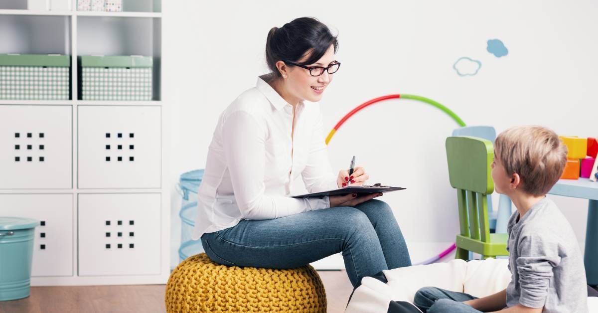 A young boy sits on a beanbag chair while looking up at a young woman who is holding a clipboard and smiling back at him.