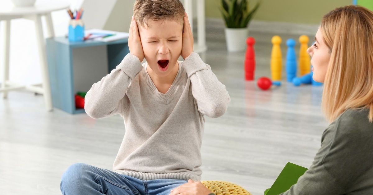 A woman sits on the ground next to a young boy showing distress with both hands covering his ears.