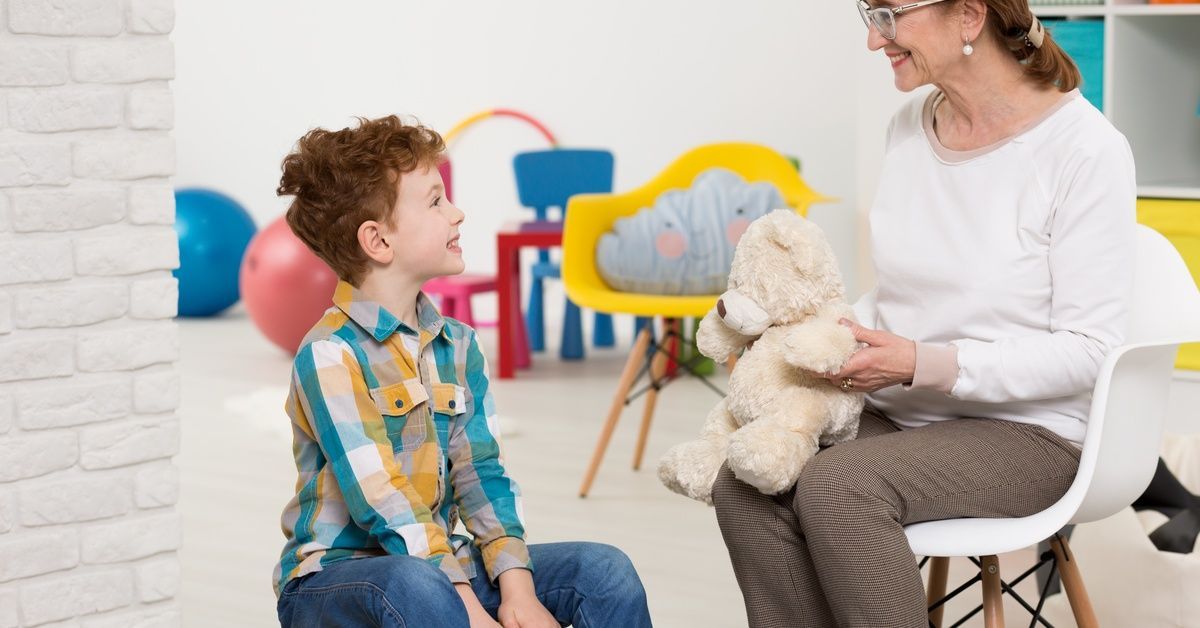 A woman with glasses sits in a chair and holds a stuffed bear while smiling down at a young boy sitting on a beanbag.