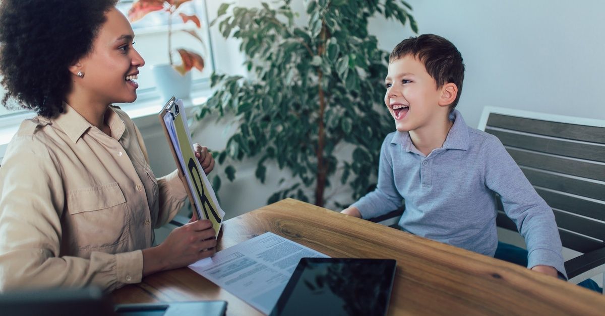 A little boy sits at a table while smiling at the woman across from him who holds up a clipboard showing the letter 'R'.