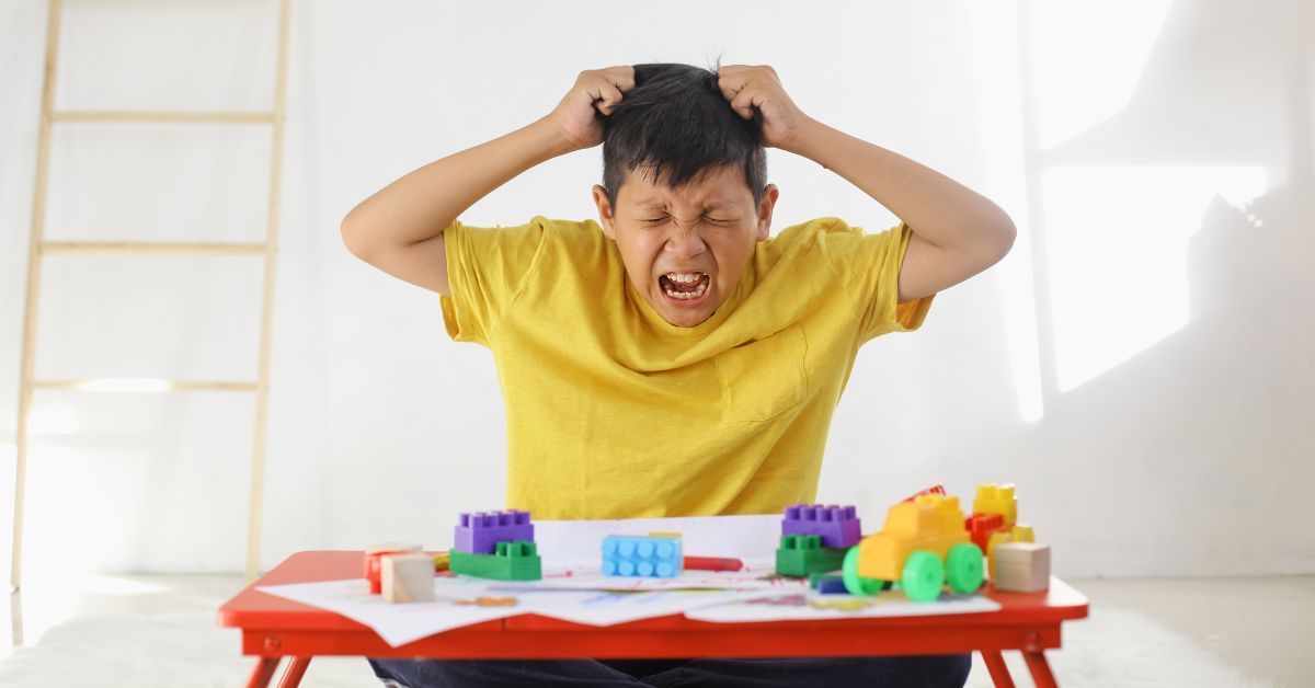 A little boy wearing a yellow shirt sits at a desk covered in blocks while pulling his hair and closing his eyes.