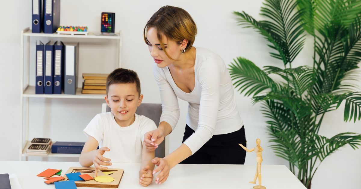 A little boy sits at a desk while playing with multi-colored puzzle pieces. The woman next to him helps him.