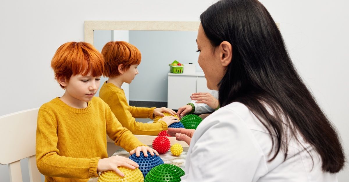 A woman wearing white sits at a table next to a young boy who is touching half balls to work on sensory exposure.