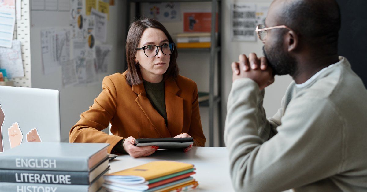 A man speaking with the woman across from him, who is sitting at a desk covered in school subject textbooks.