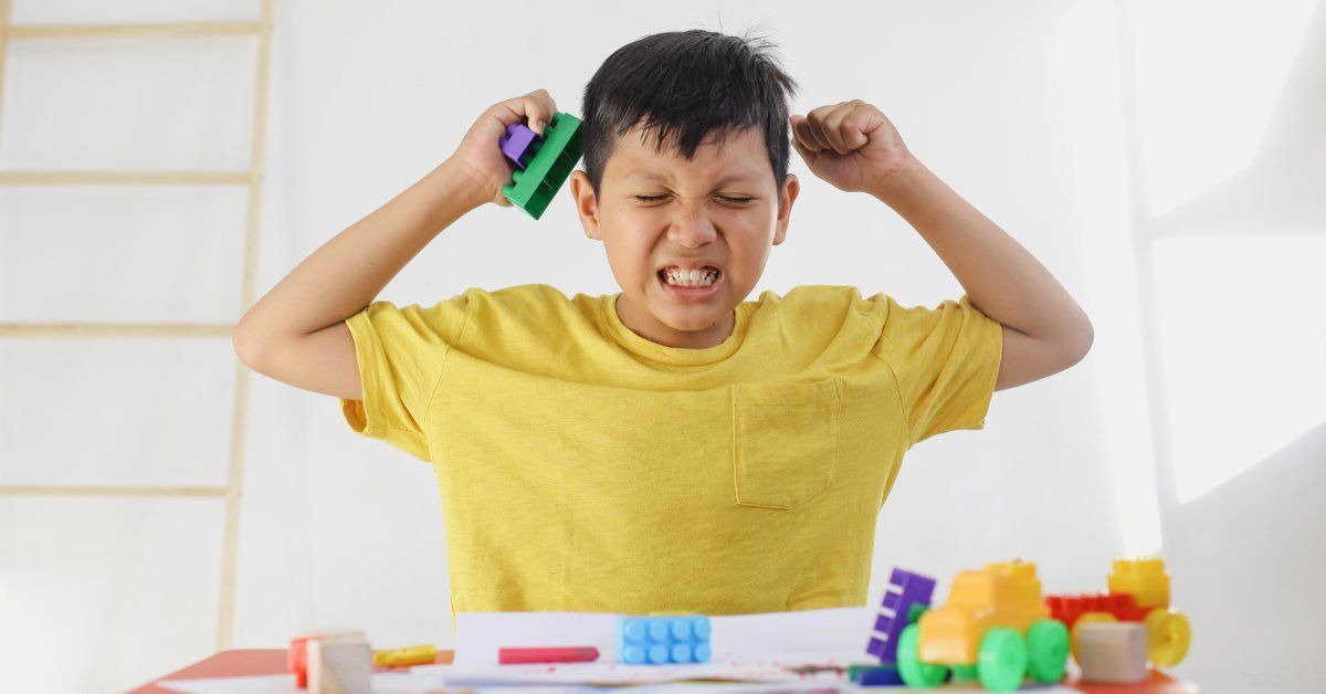 A little boy wearing a yellow shirt sits at a table while making an angry face and holding toys close to his head.