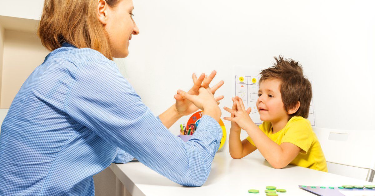 A little boy leans up against a table with his hands together, mirroring the woman sitting across from him.