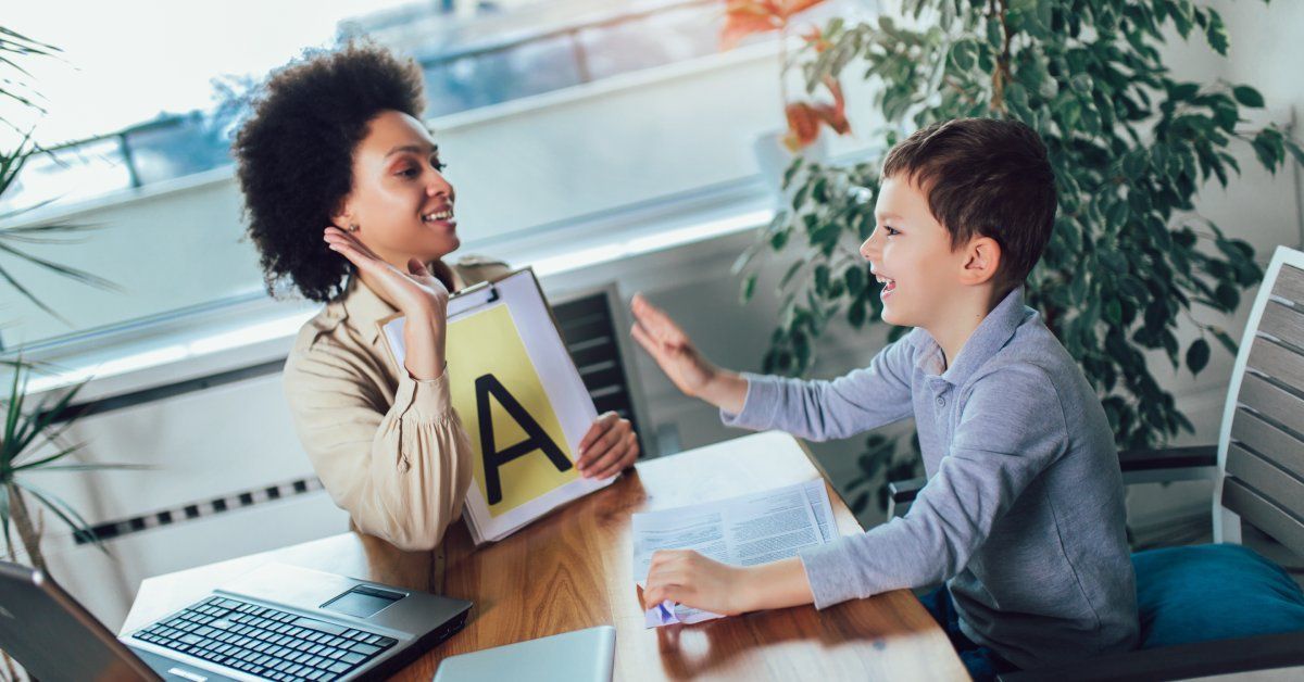 A woman sitting at a desk next to a smiling young boy, while holding a clipboard that has the letter