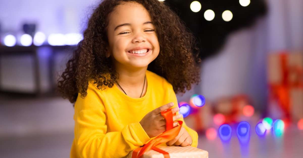 A happy girl in a sweater is unwrapping her Xmas gift and smiling at home.