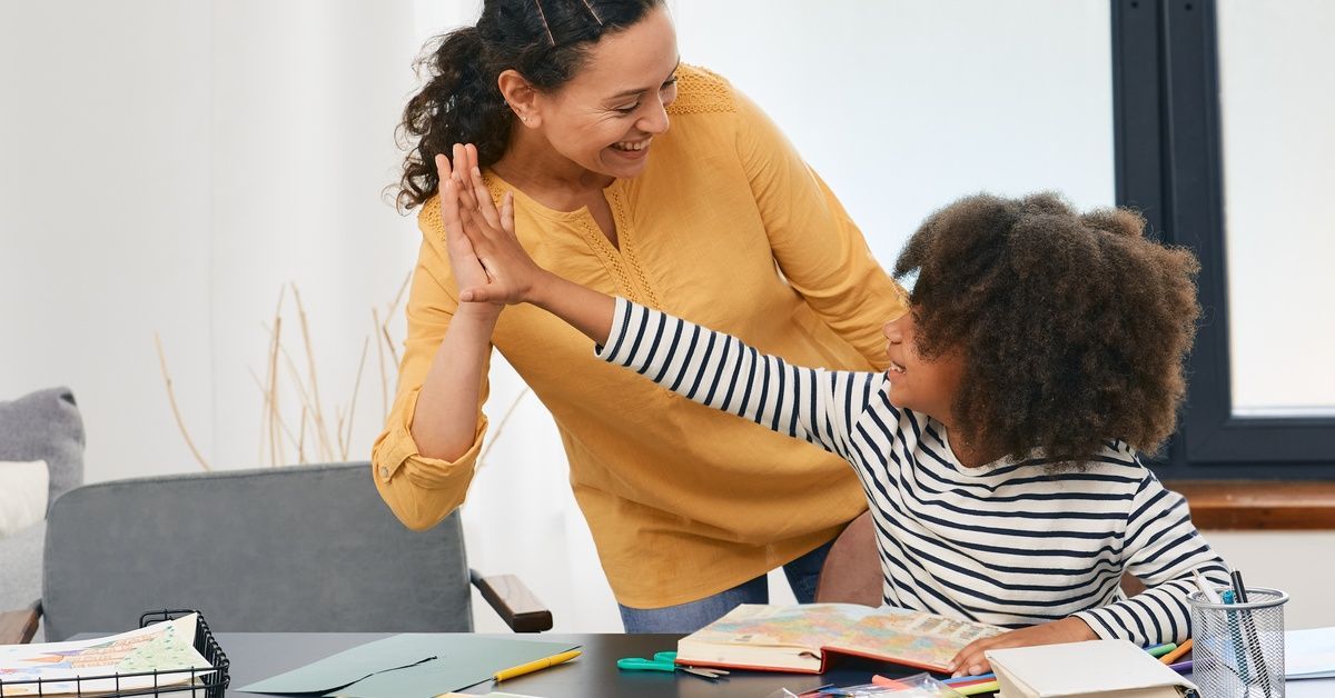 A woman in a yellow top and a child in a striped shirt share a joyful high-five over a desk filled with school supplies.