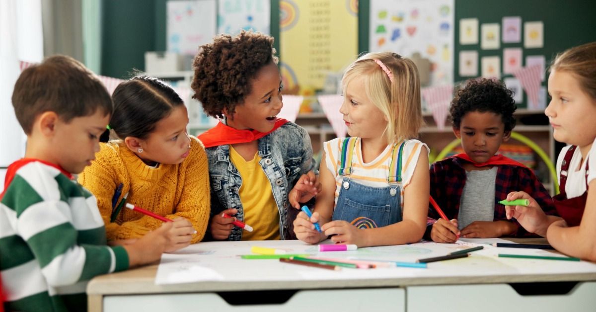 Six children sit at a classroom table, drawing with colorful markers and talking to each other about their drawings.